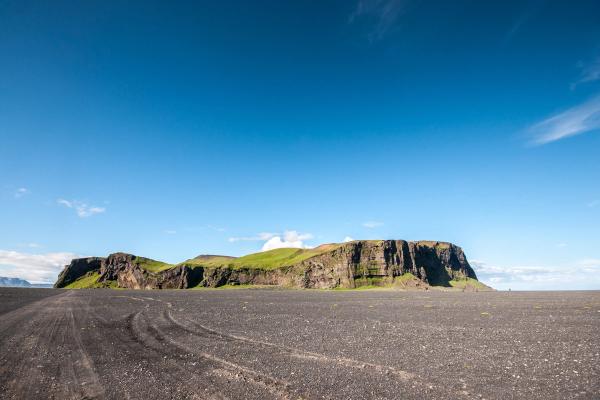 Vast black volcanic plain with tire tracks, leading to a long, green-topped cliff under a bright blue sky.