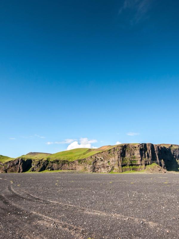 A wide black volcanic plain with tire tracks leads to a long, green-topped mesa under a bright blue sky.