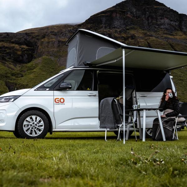 Una furgoneta Go Camper blanca con techo elevable y toldo extendido, una mujer sentada debajo bebiendo en un paisaje montañoso y herboso.
