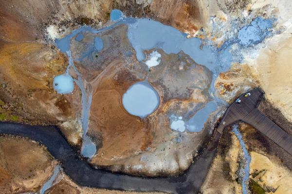 Aerial view of a geothermal area with pale blue pools and streams, surrounded by colorful orange and brown earth, and a wooden boardwalk.