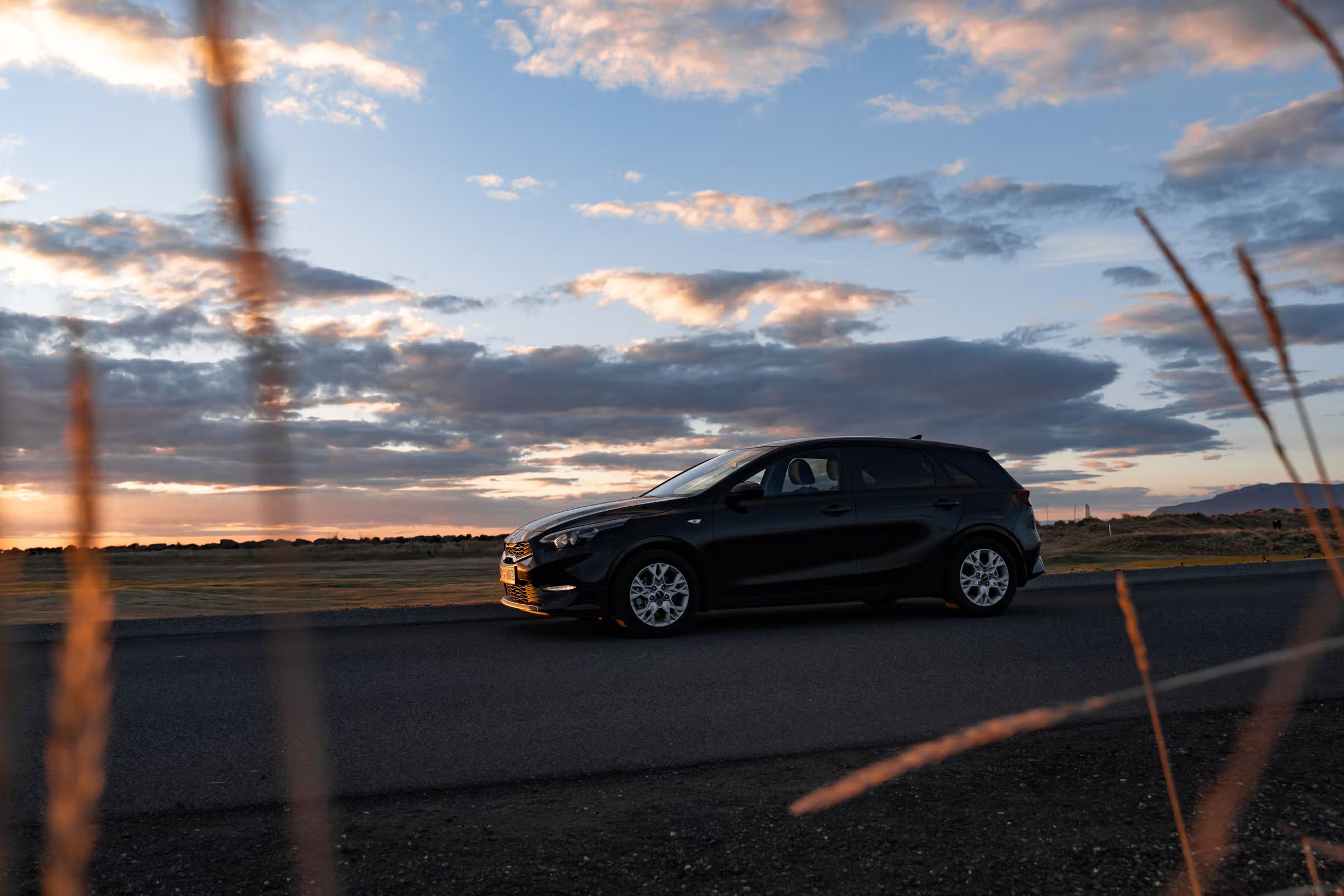 Un coche negro aparcado en una carretera, con hierba alta en primer plano y un cielo al atardecer lleno de nubes de colores.
