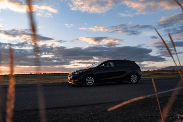A black car parked on a road with tall grass in the foreground, under a sunset sky with colorful clouds.