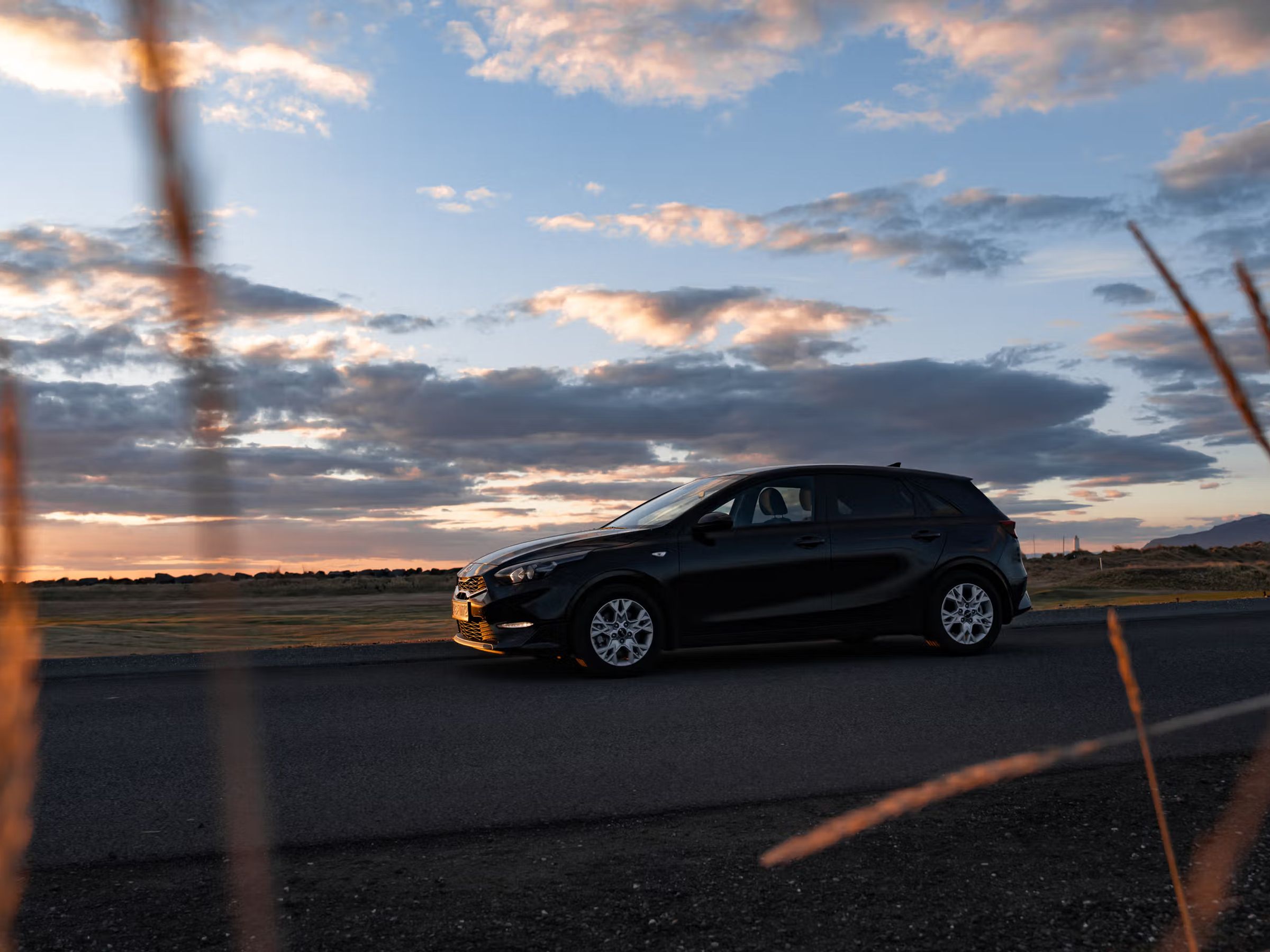 A black car parked on a road at sunset, with blurry tall grass in the foreground.