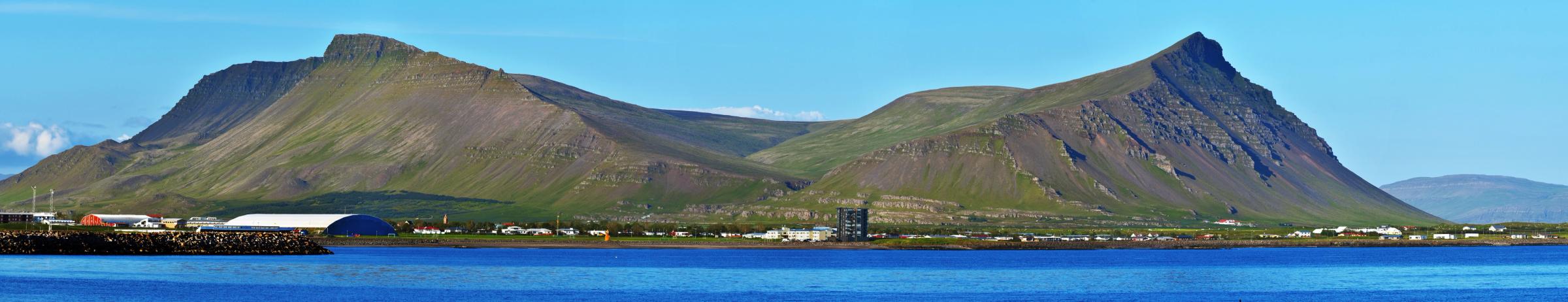 Panoramic view of a town on a blue bay with large, green-sloped mountains under a clear sky.