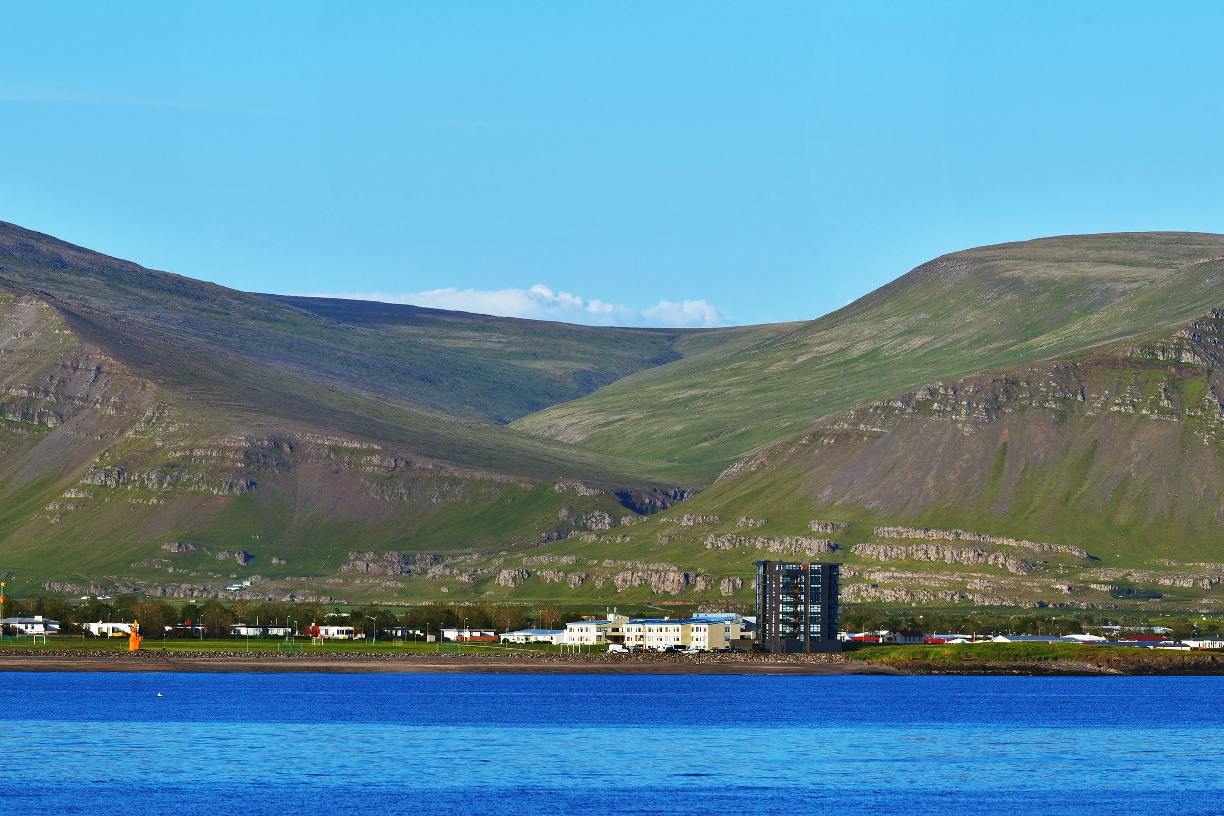 Panoramic view of a town on a blue bay with large, green-sloped mountains under a clear sky.
