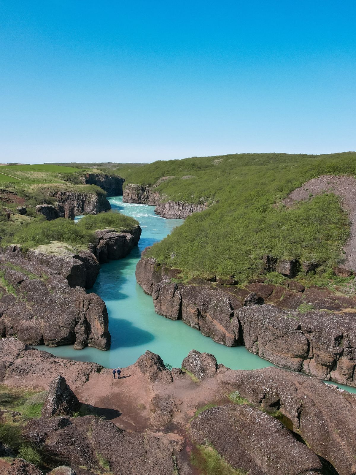 Un río de color turquesa brillante fluye a través de un cañón rocoso con vegetación verde bajo un cielo azul claro.