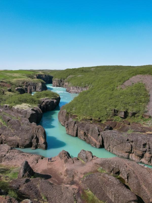 A bright turquoise river flows through a rocky canyon with green vegetation under a clear blue sky.