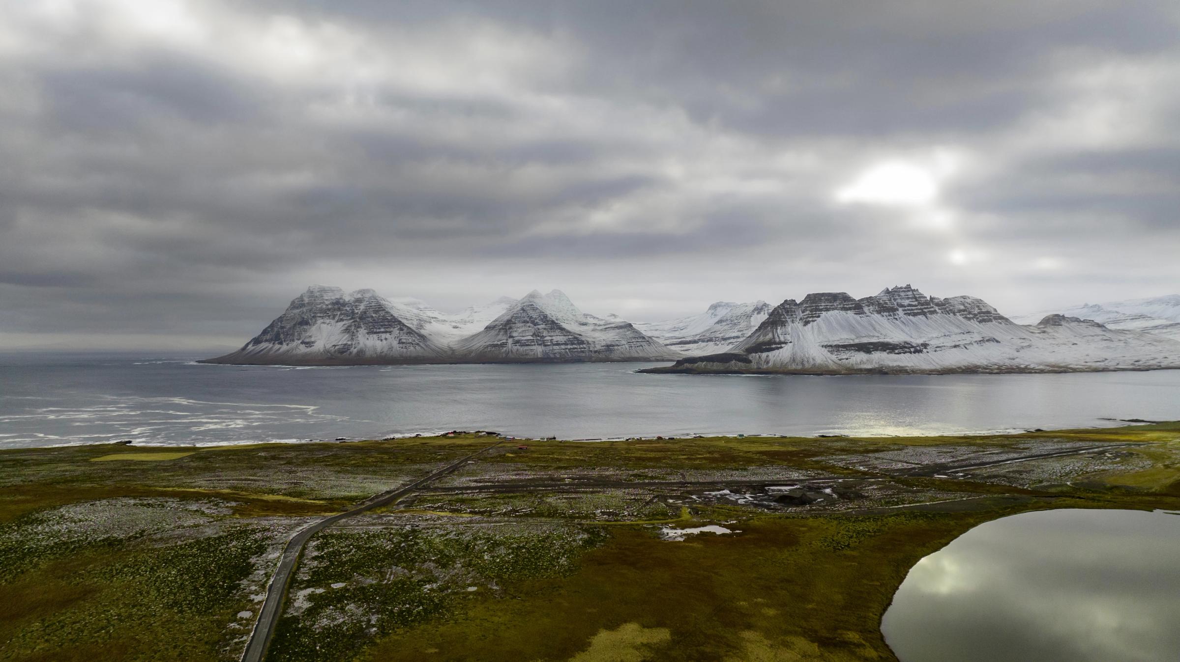 Snowy mountains rise across a bay from a green-brown landscape with a winding road and a small pond, all under a cloudy sky.