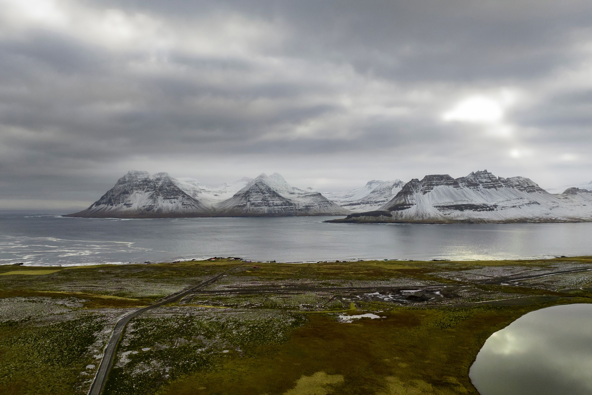 Snowy mountains rise across a bay from a green-brown landscape with a winding road and a small pond, all under a cloudy sky.