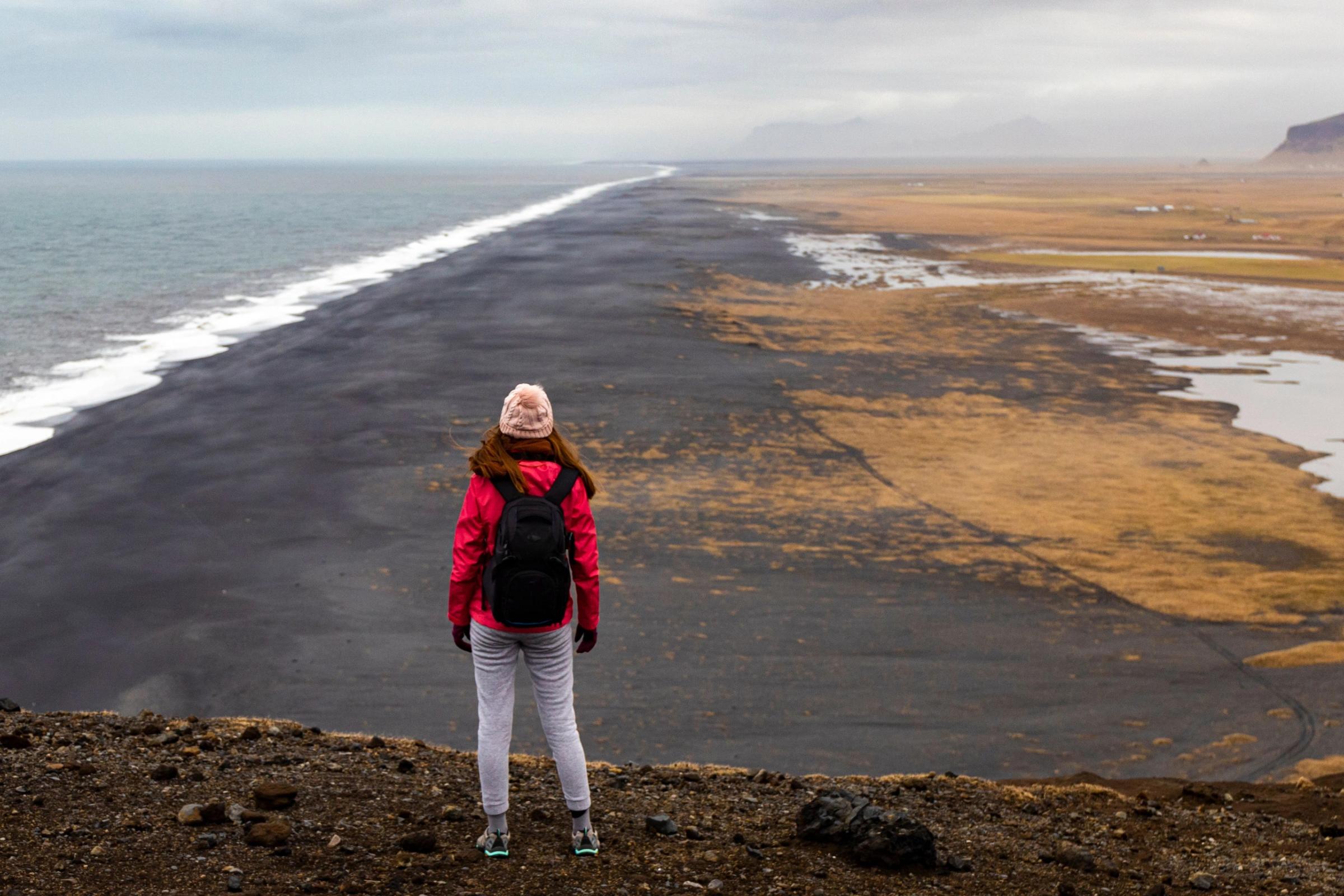 Girl in red jacket and backpack looks at a vast empty Dyrhólaey black sand beach