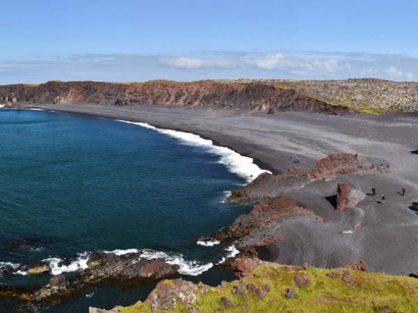 A panoramic view of a dark volcanic sand beach bordering a blue ocean, framed by rugged, moss-covered cliffs under a clear sky.