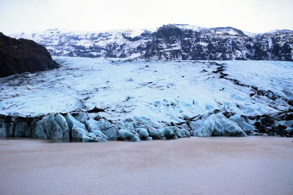 A vast blue-white glacier descends into a murky glacial lake, with snow-capped mountains in the background.