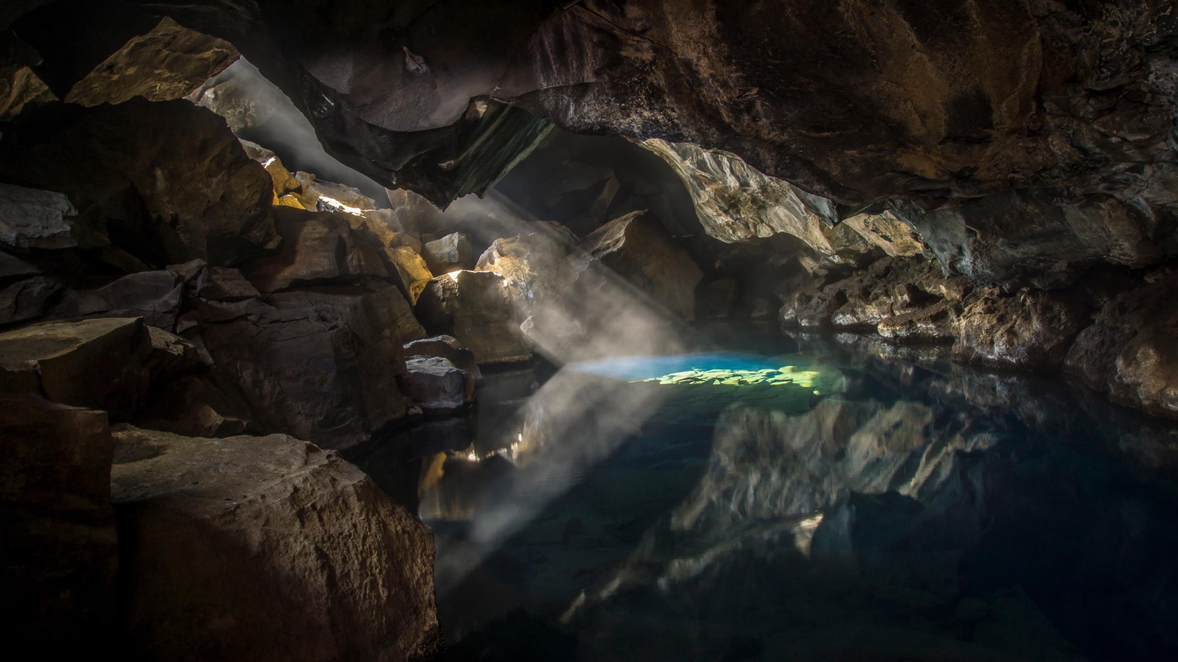 La luz del sol se cuela en una cueva oscura, iluminando la neblina sobre una poza de agua turquesa que refleja todo como un espejo.