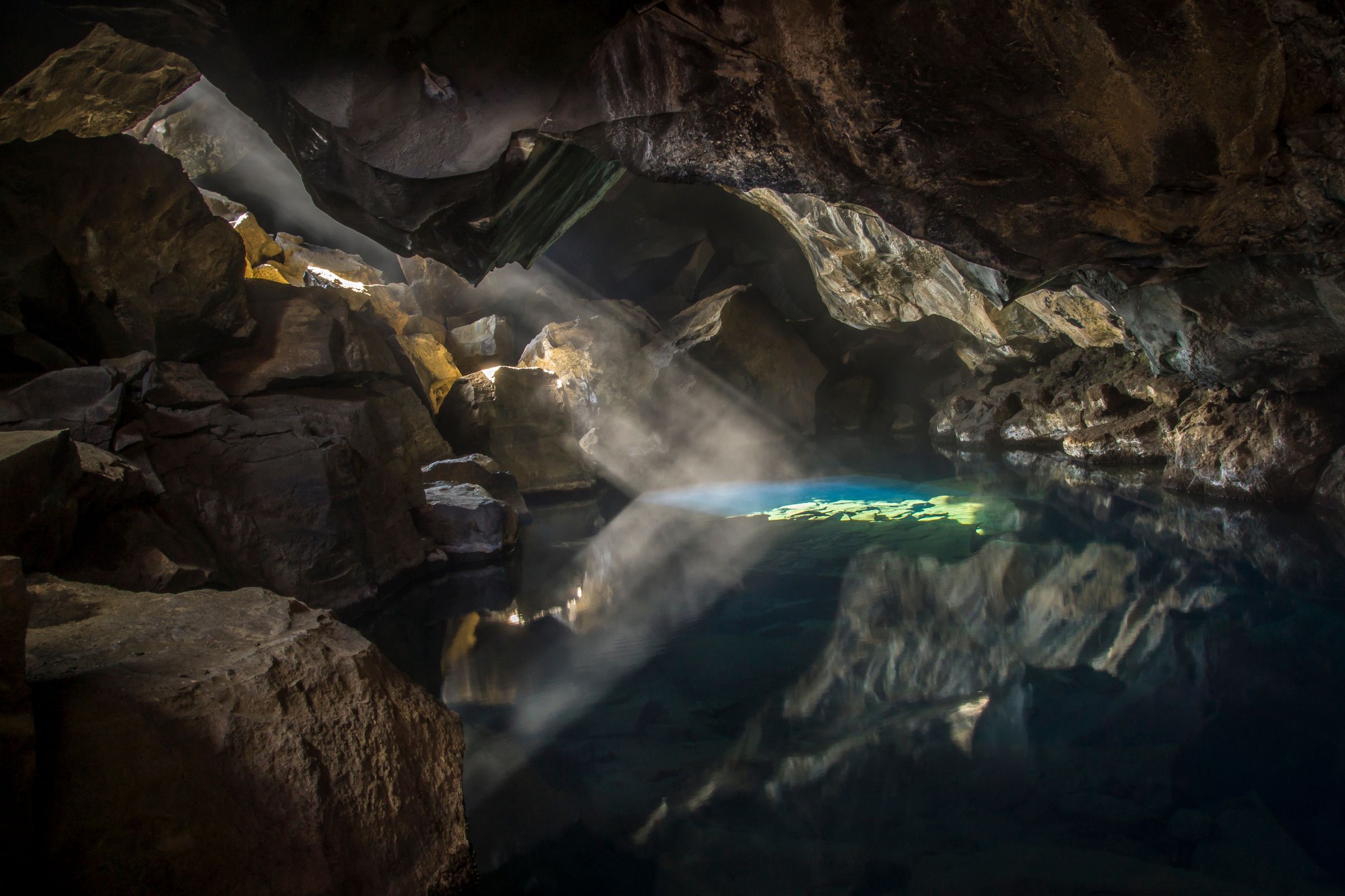 La luz del sol se cuela en una cueva oscura, iluminando la neblina sobre una poza de agua turquesa que refleja todo como un espejo.