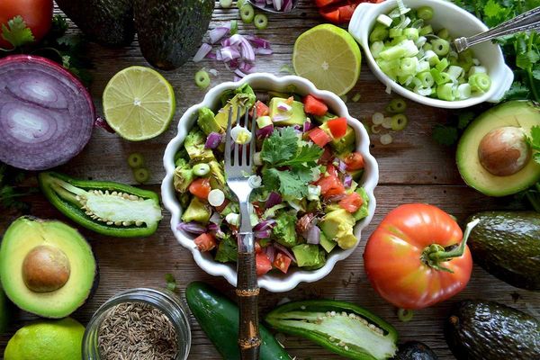 A bowl of avocado and tomato salad with a fork, surrounded by fresh produce on a rustic wooden table.