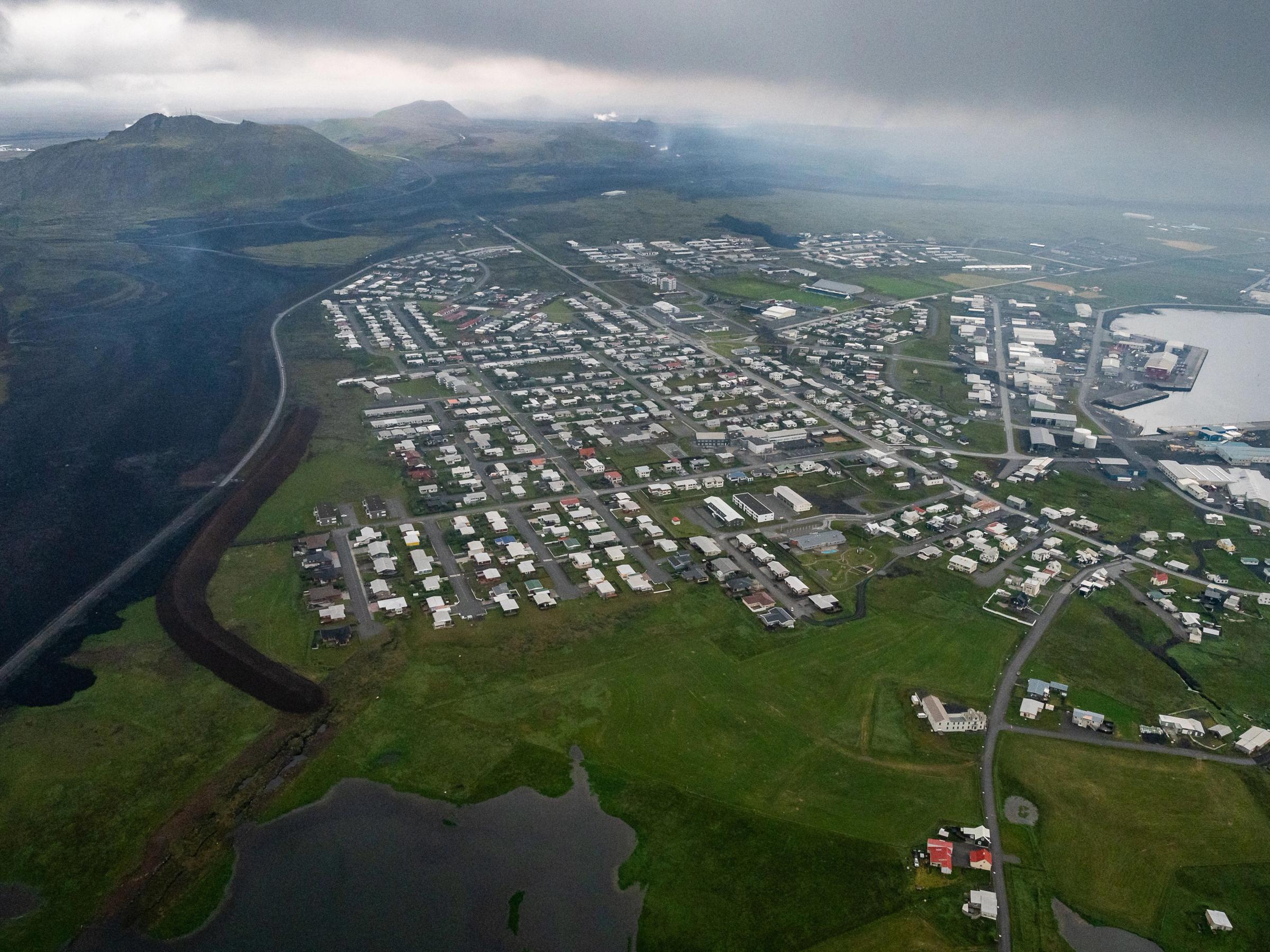 Aerial view of a town nestled between a dark lava field, green landscape, and a harbor under an overcast sky.