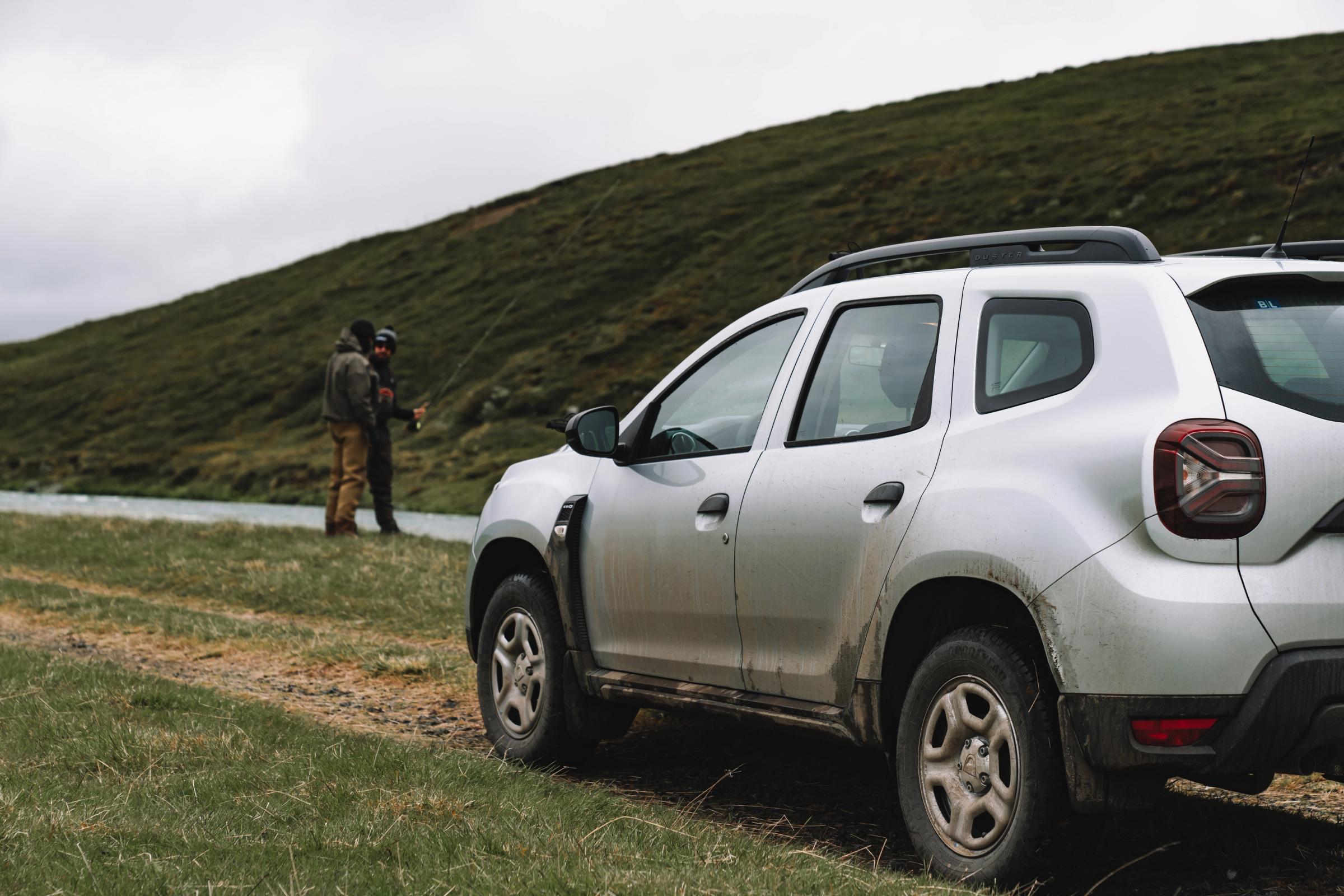 Dacia Duster rear quarter view parked on grass beside a river with a person fishing
