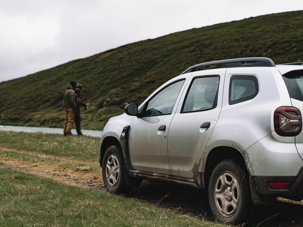 Vista trasera del Dacia Duster Riverside Vista trasera de tres cuartos de un Dacia Duster aparcado sobre la hierba junto a un río, con una persona pescando
