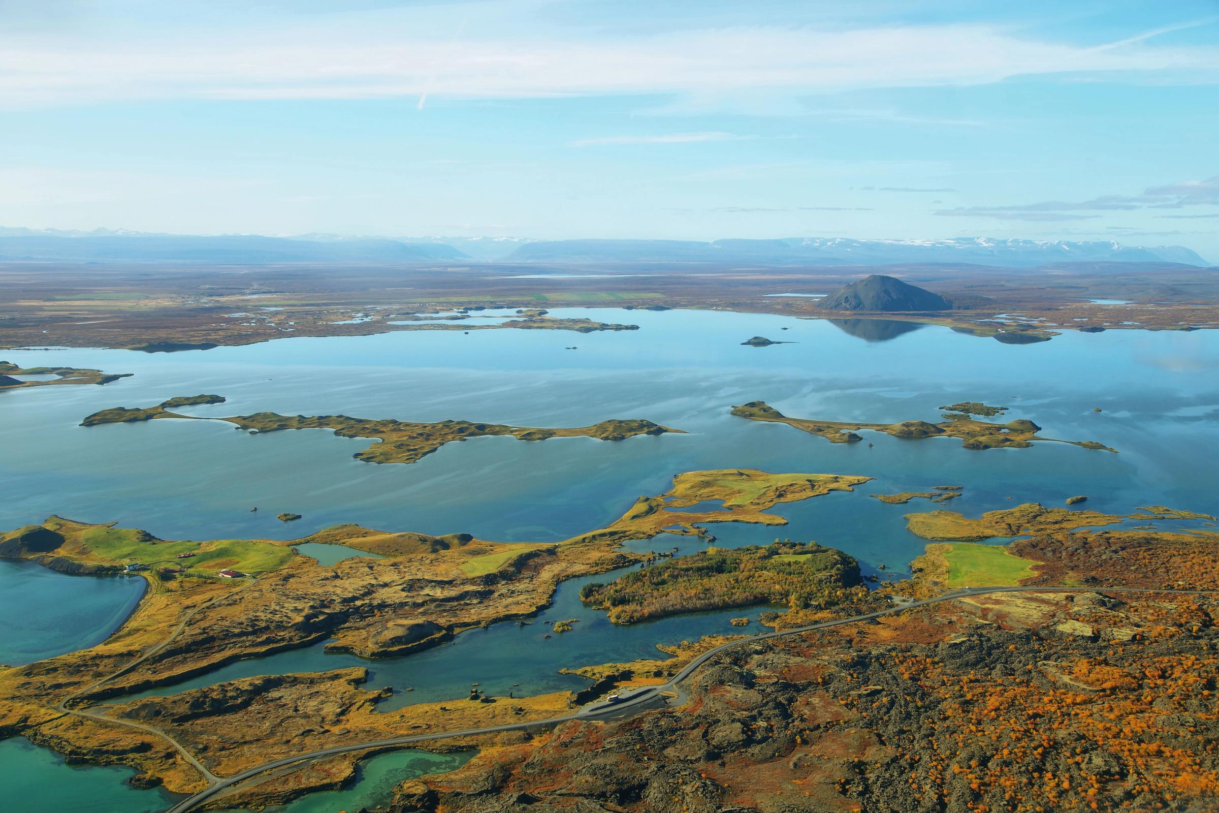 Aerial view of a lake with numerous islands, a conical hill reflecting in the distance, and surrounding autumnal terrain.