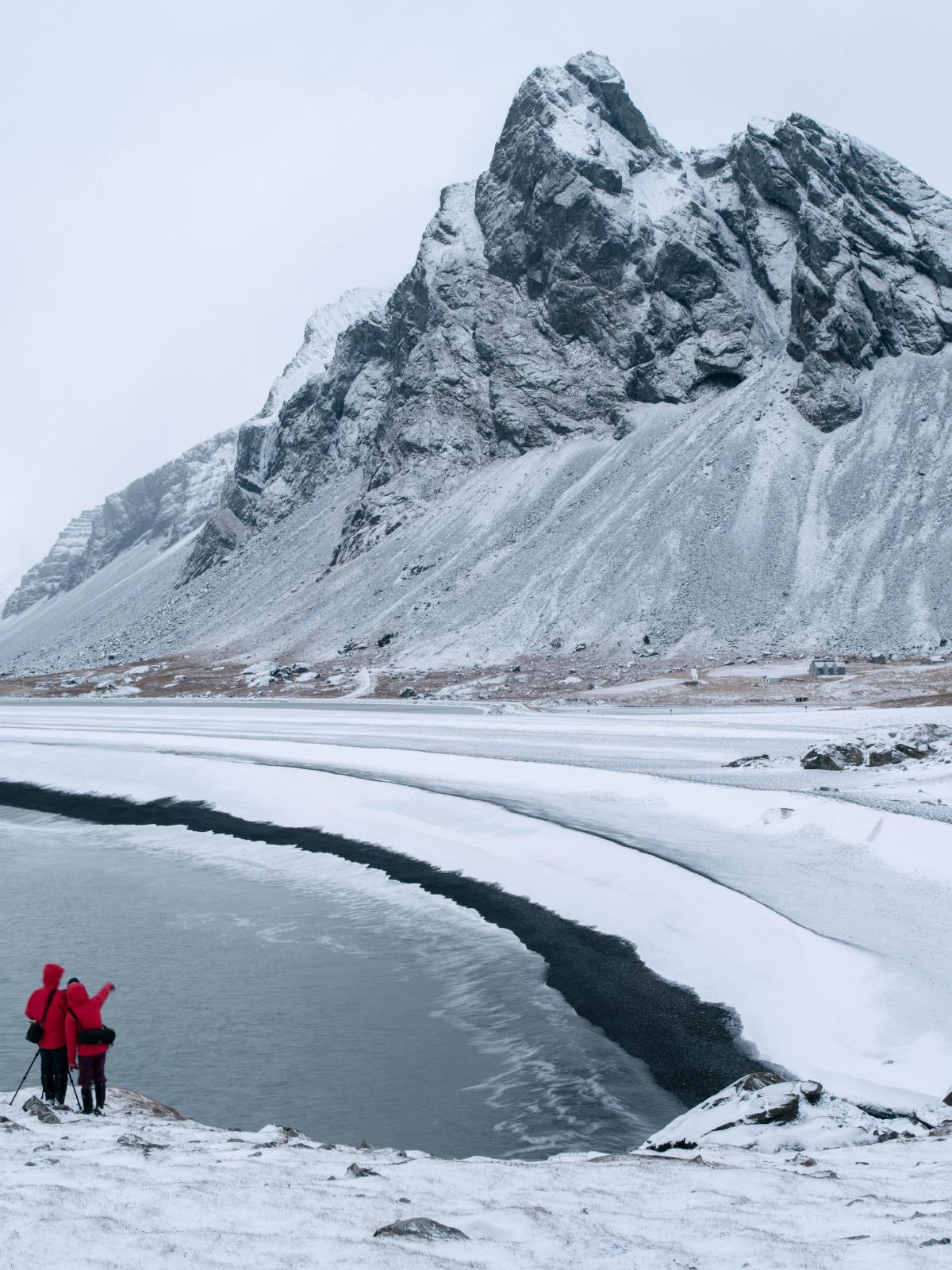Two people in red jackets stand on a snowy bank by a partially frozen lake, framed by snow-capped mountains.