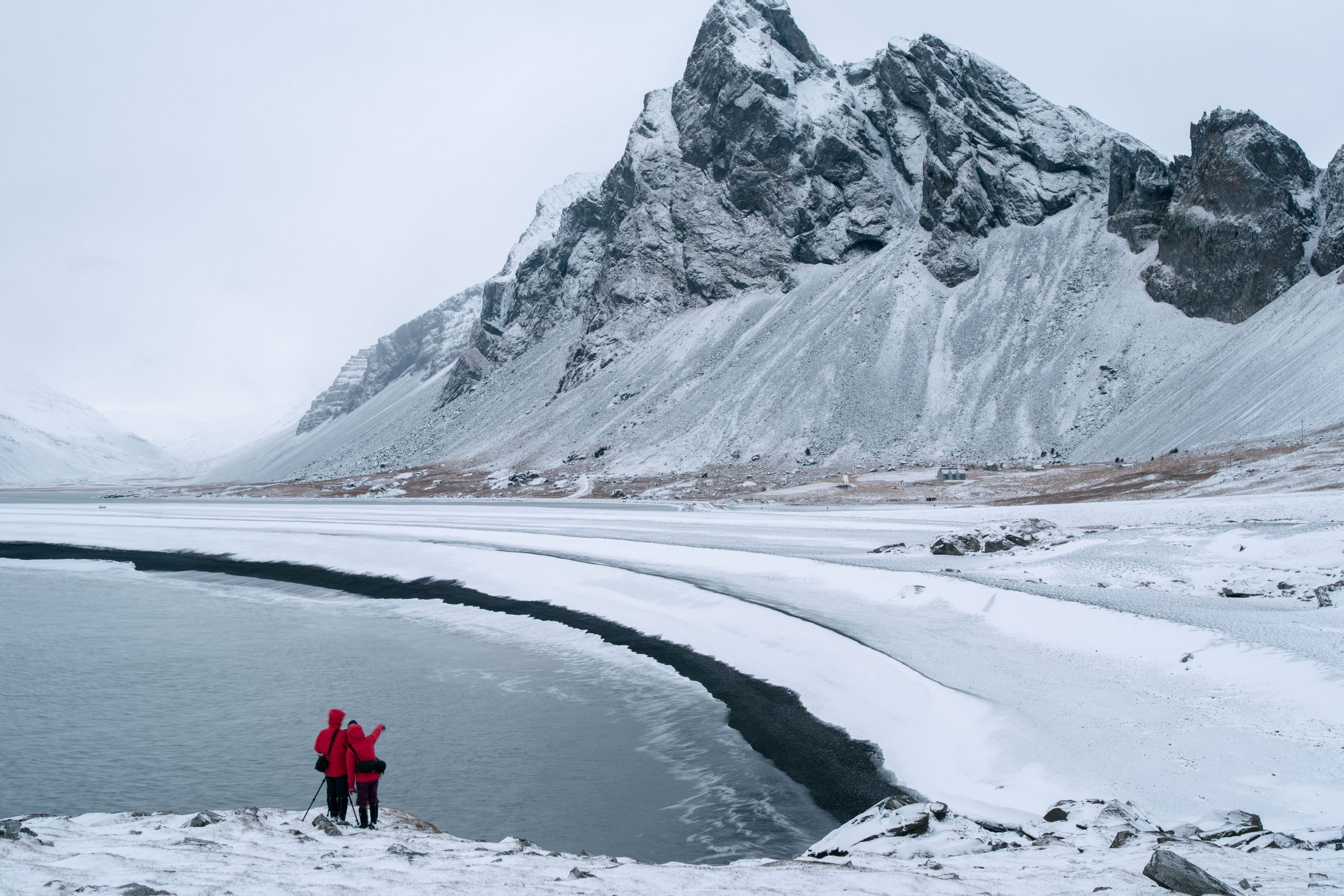 Dos personas con chaquetas rojas están de pie en una orilla nevada mirando un lago parcialmente congelado y montañas cubiertas de nieve.