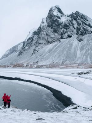 Dos personas con chaquetas rojas están de pie en la orilla nevada junto a un lago parcialmente congelado, enmarcados por montañas cubiertas de nieve.