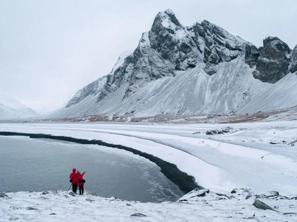 Dos personas con chaquetas rojas están de pie junto a un lago parcialmente congelado, admirando las montañas cubiertas de nieve.
