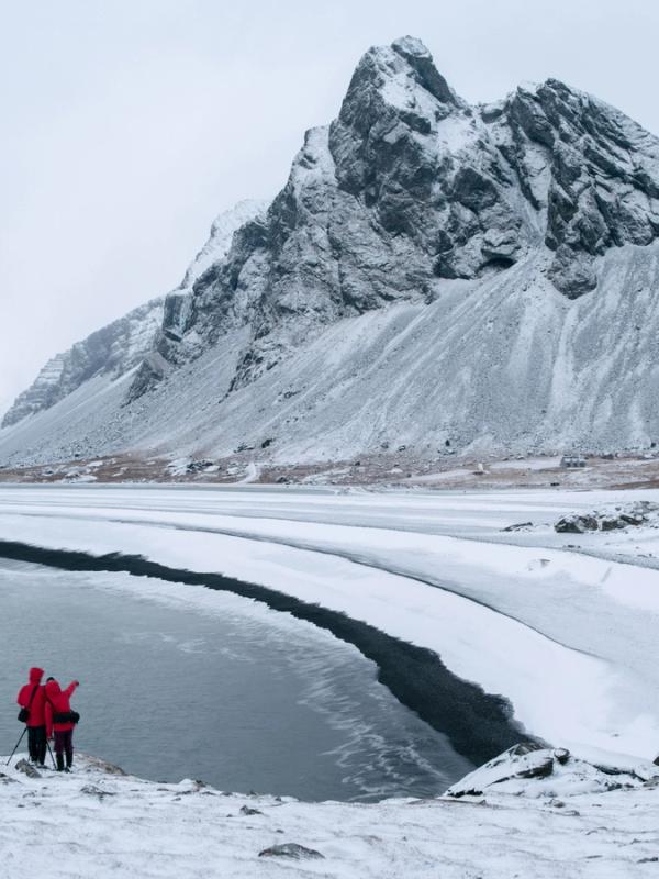 Two people in red jackets stand on a snowy bank by a partially frozen lake, framed by snow-capped mountains.