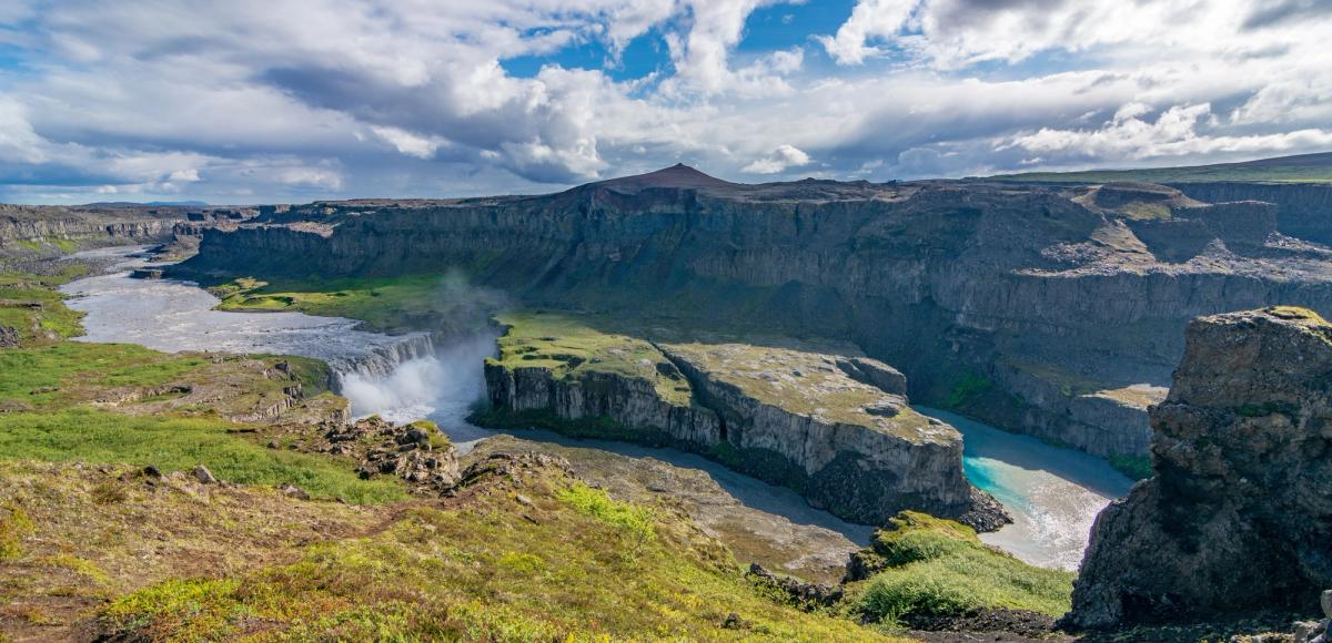 A powerful waterfall cascading into a river flowing through a dramatic, green canyon under a partly cloudy sky.