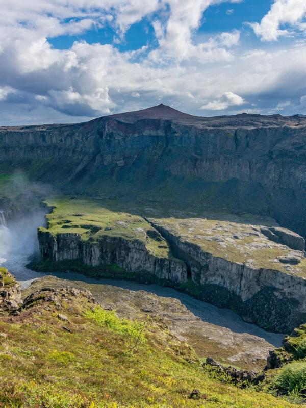 A powerful waterfall cascading into a river flowing through a dramatic, green canyon under a partly cloudy sky.