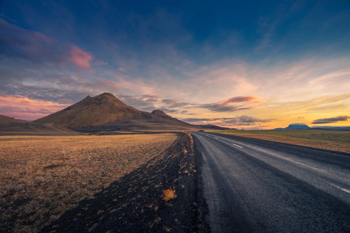 A road leads through dry fields towards mountains under a vibrant sunset sky.