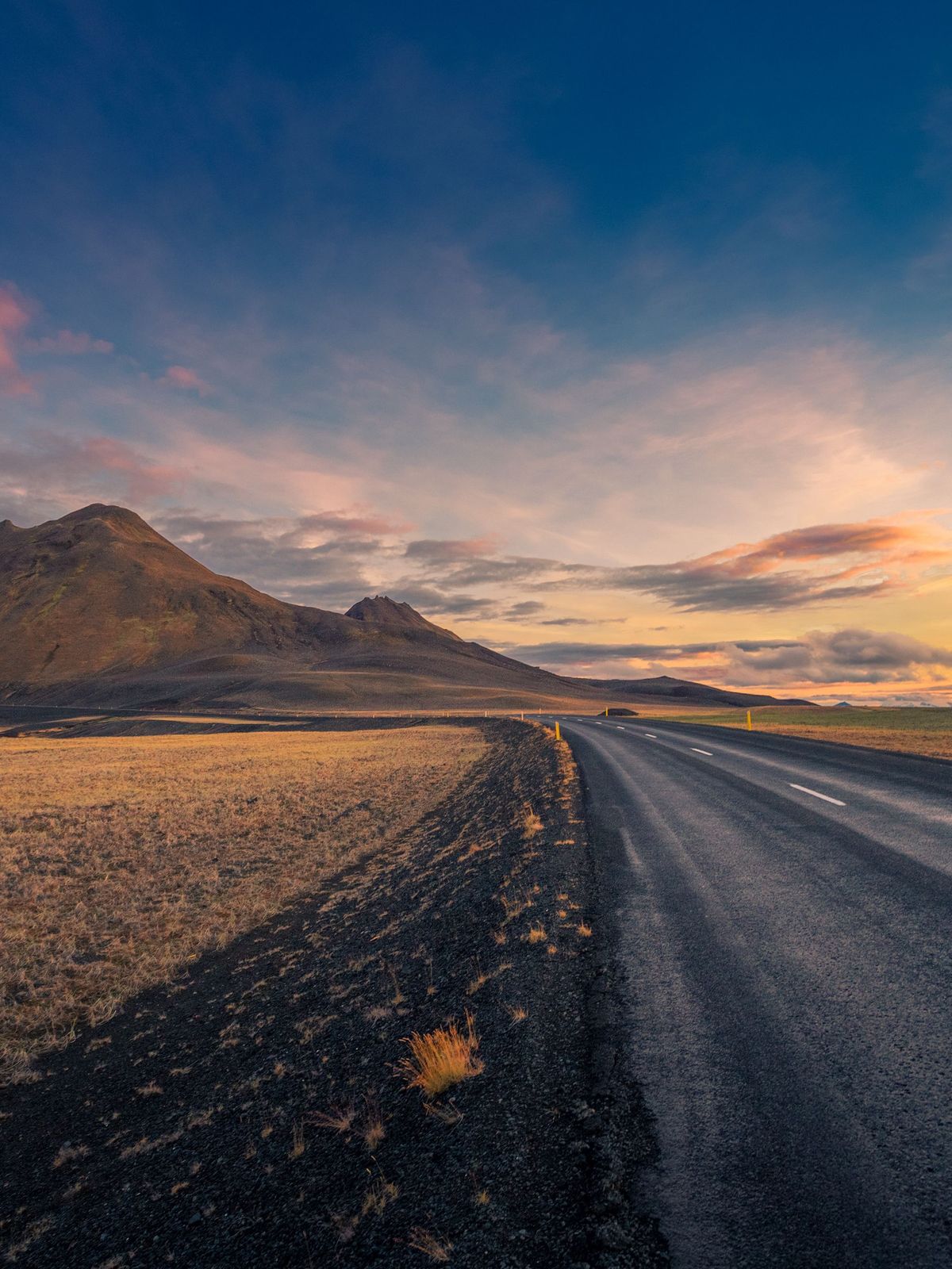 A road leads through dry fields towards mountains under a vibrant sunset sky.