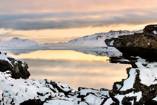 Snowy rocks frame a calm lake reflecting a warm, cloudy sky with distant snow-capped mountains.