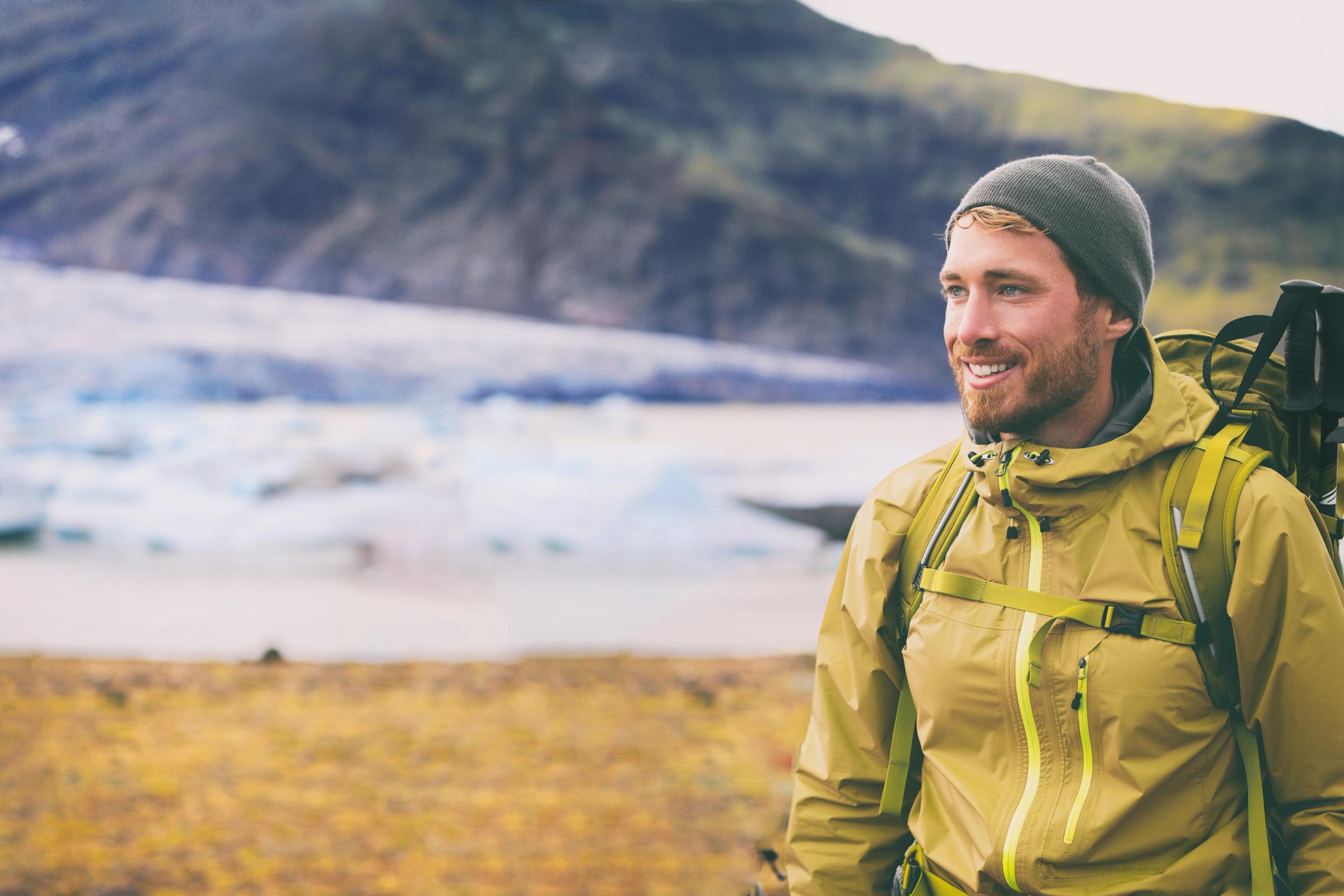 A smiling man in an olive green hiking jacket and beanie stands in front of a glacier and mountains.