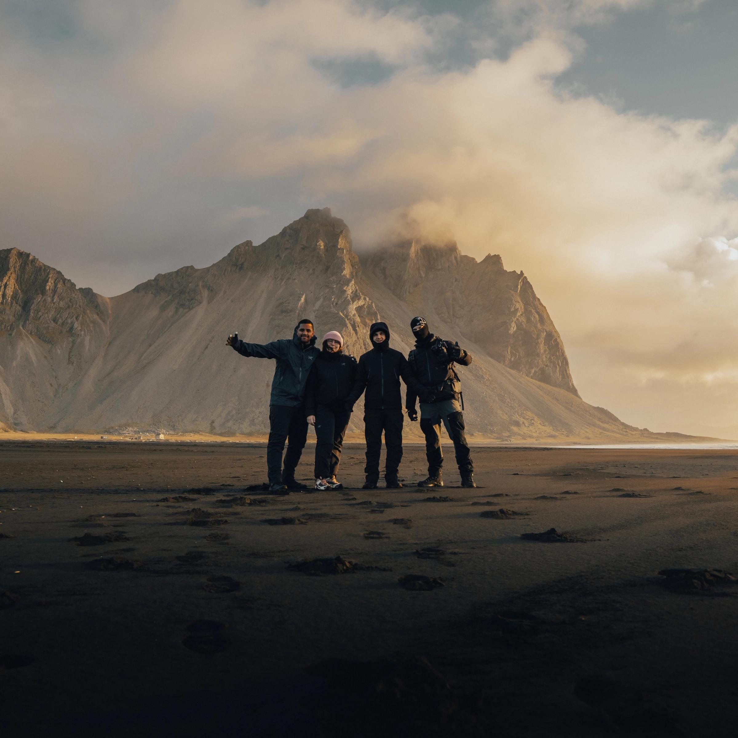 Four people stand on a black sand beach in front of large, rugged mountains under a golden, cloudy sky.