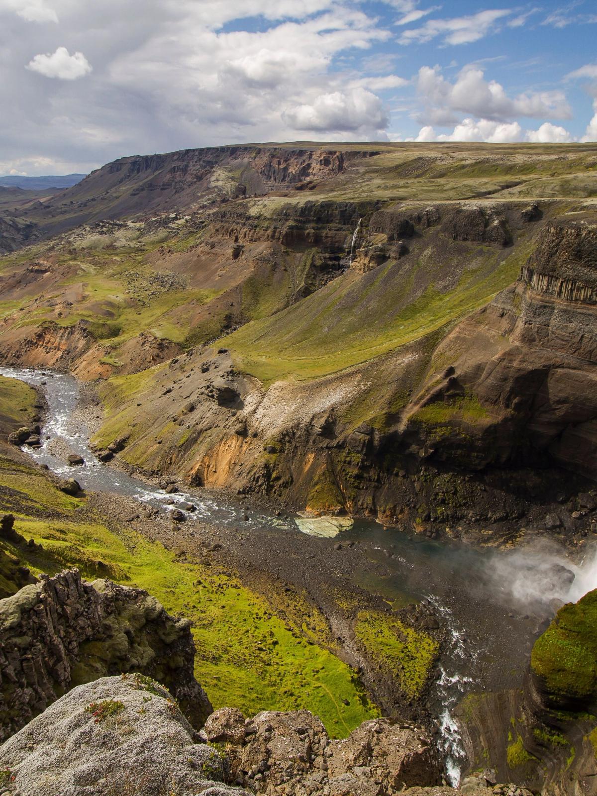 A powerful waterfall plunges into a river winding through a vast green and rocky canyon.