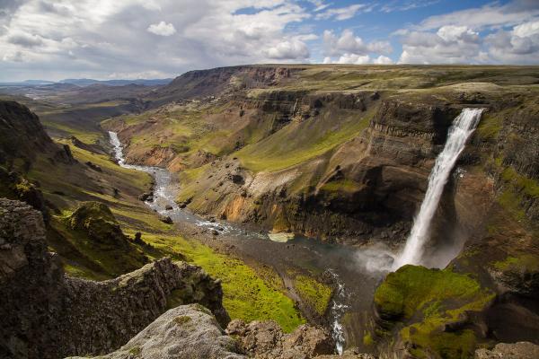 A large waterfall cascades into a green and rocky canyon with a winding river under a cloudy sky.