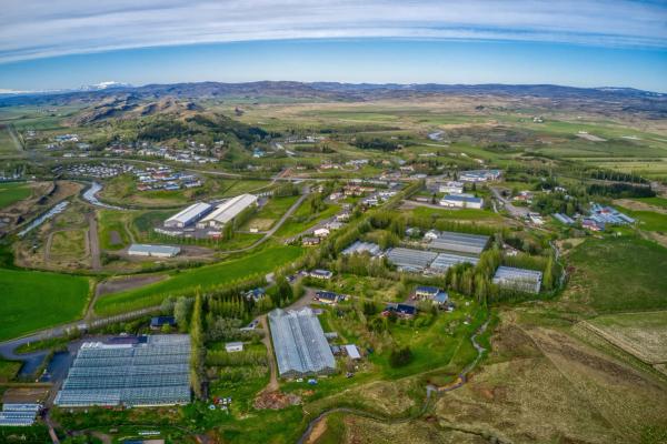 Aerial view of a town nestled among green fields and numerous greenhouses, with a river winding through and snow-capped mountains in the background.