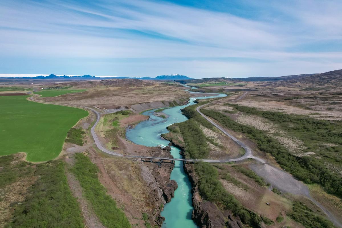 Aerial view of a bright turquoise river winding through a valley with green fields, roads, and distant mountains.