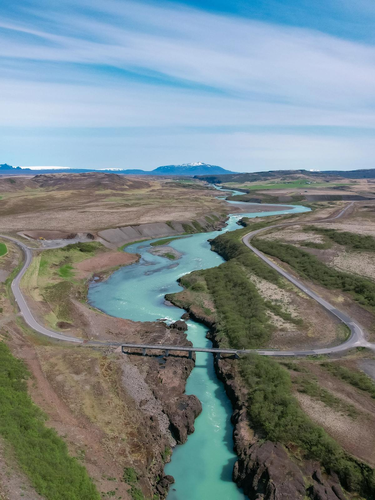 Aerial view of a bright turquoise river winding through a valley with green fields, roads, and distant mountains.