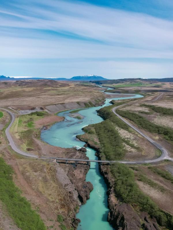 Aerial view of a bright turquoise river winding through a valley with green fields, roads, and distant mountains.