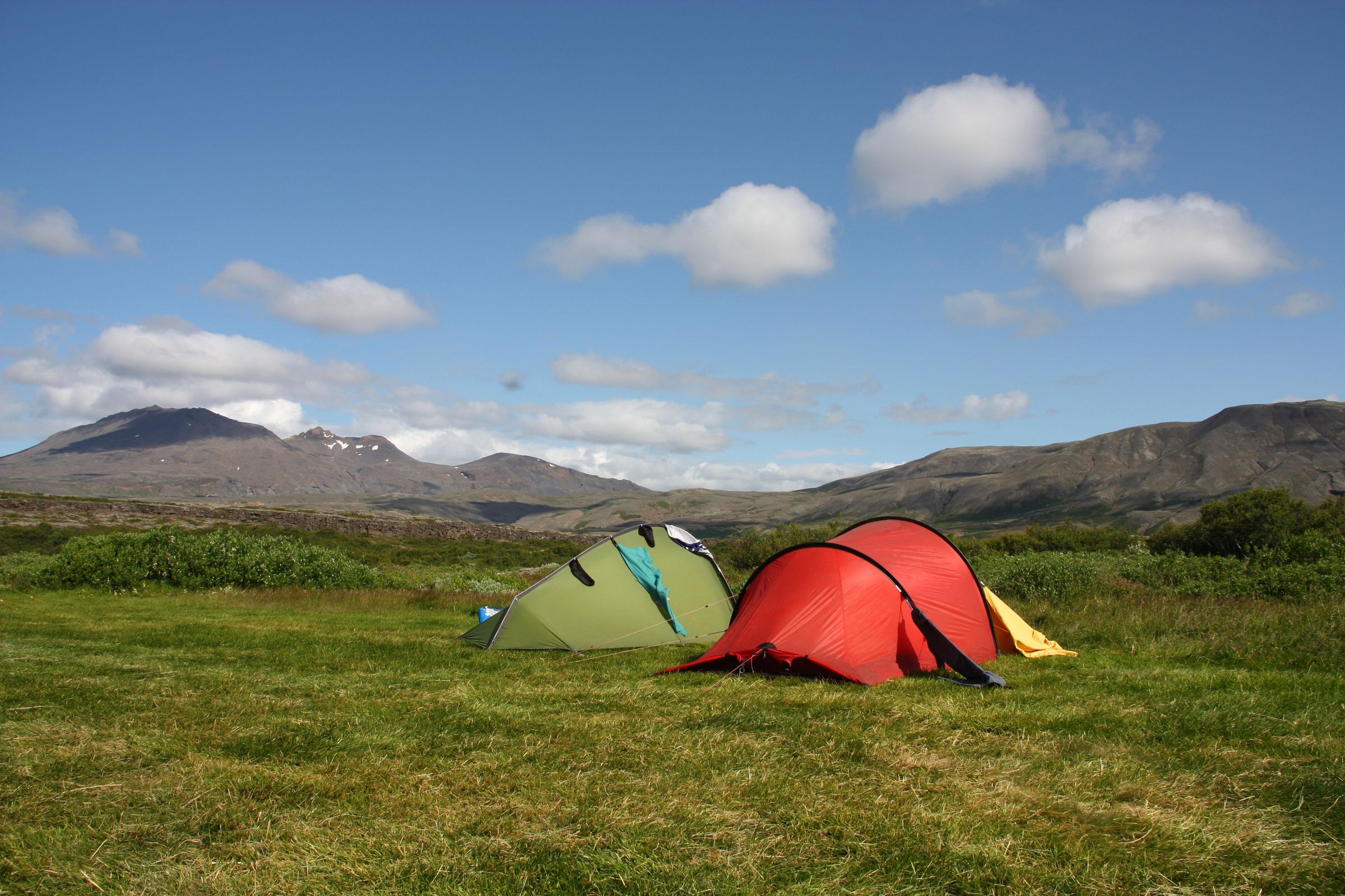 Two tents, one green and one red, pitched on a grassy field with mountains in the background under a blue sky.