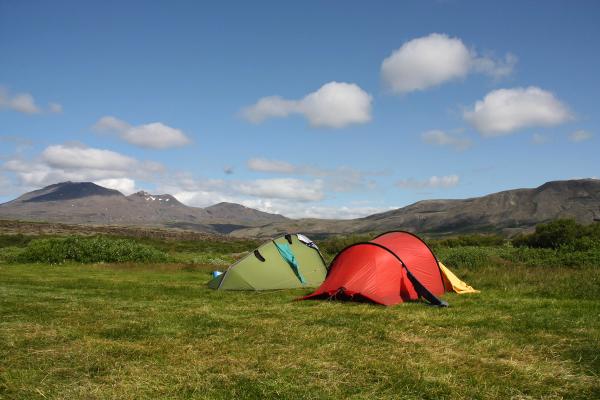 Two tents, one green and one red, pitched on a grassy field with mountains in the background under a blue sky.
