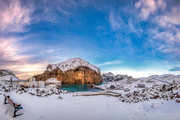 Panoramic view of a snowy, rocky coastal landscape with a large mountain, turquoise bay, industrial tanks, and a bench in the foreground under a blue and pink sky.