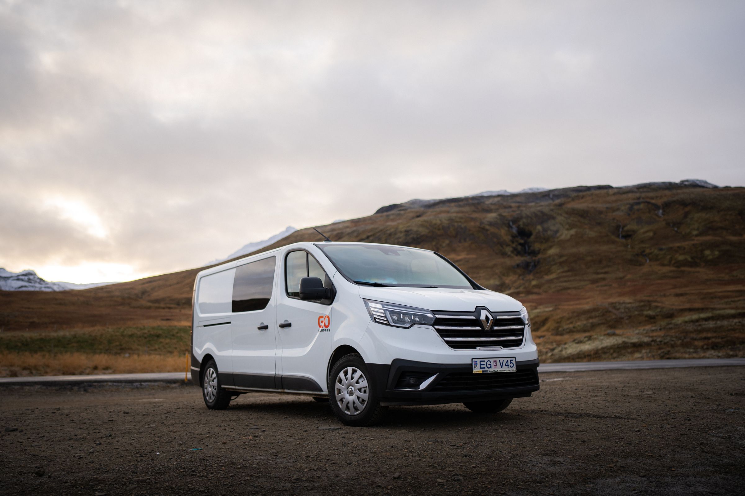 White Renault van with 'GO' logo parked on a dirt road, surrounded by mountains under a cloudy sky.