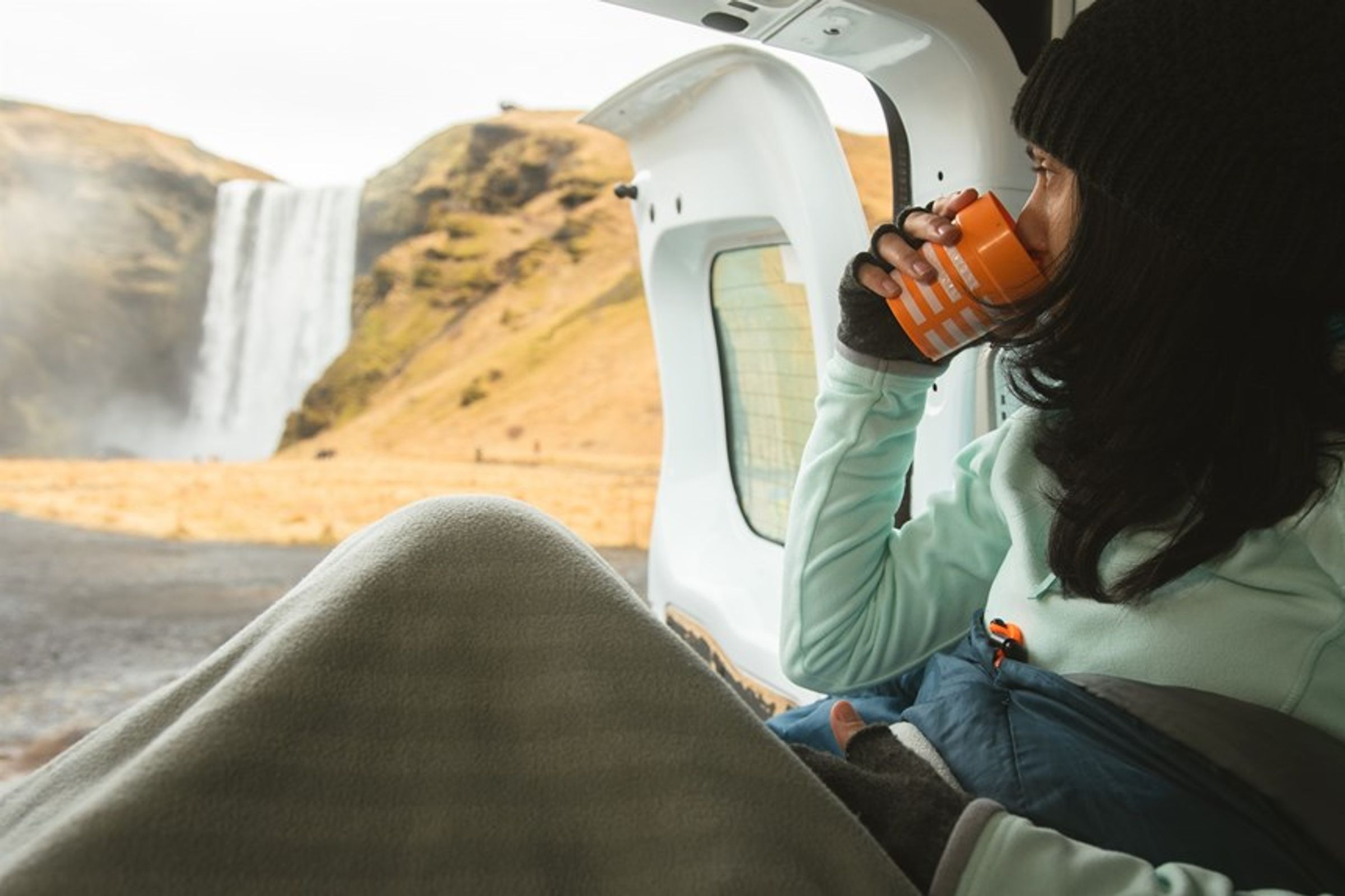 a woman is sitting in the back of a car drinking a cup of coffee by a waterfall inside a camper in iceland
