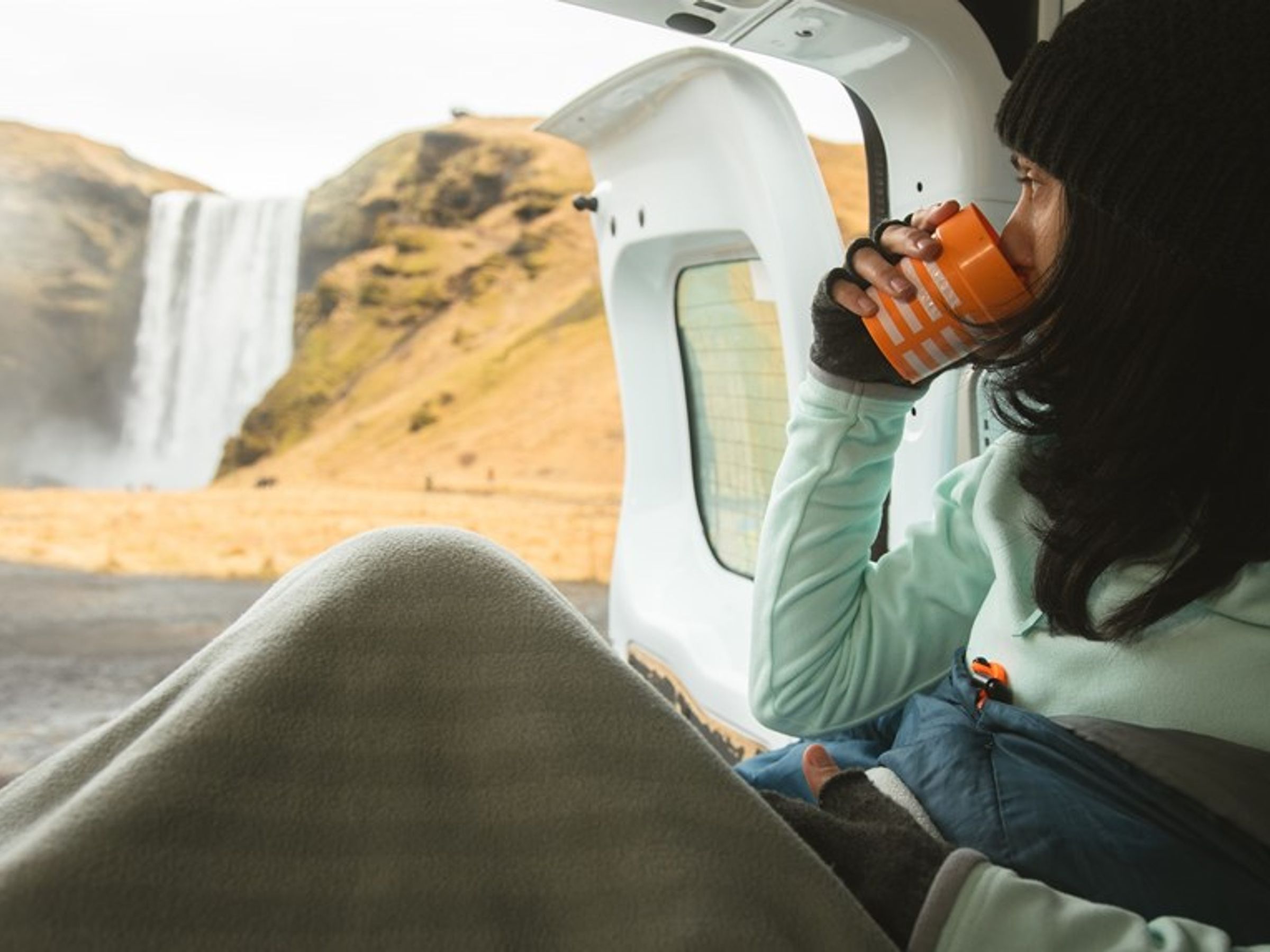 a woman is sitting in the back of a car drinking a cup of coffee by a waterfall inside a camper in iceland