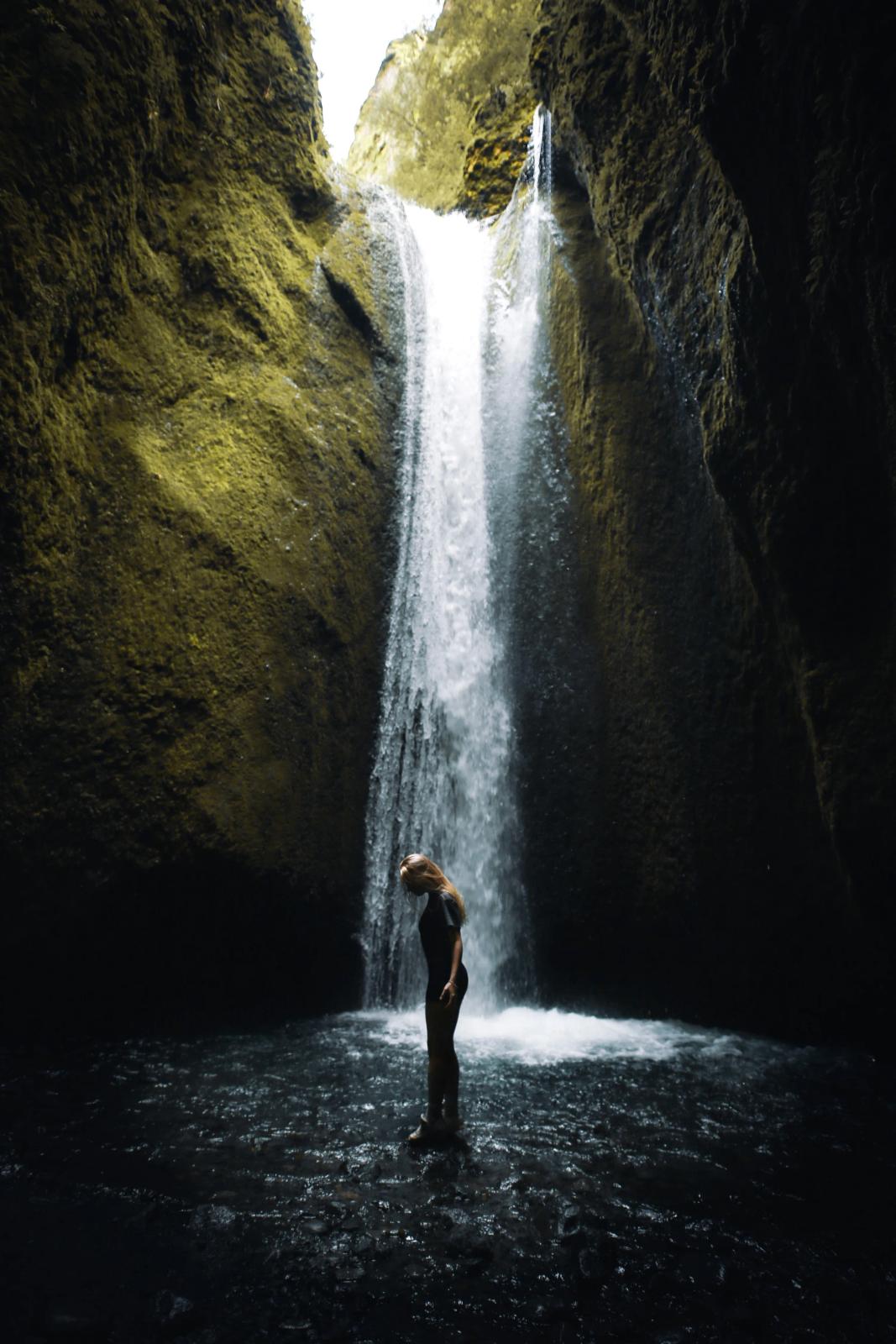 Una mujer está de pie en agua poco profunda bajo una cascada dentro de un cañón musgoso.