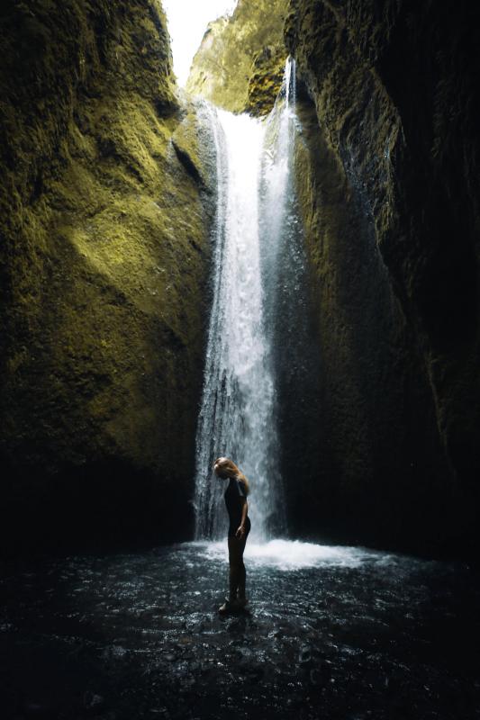 A woman stands in shallow water under a waterfall inside a mossy canyon.