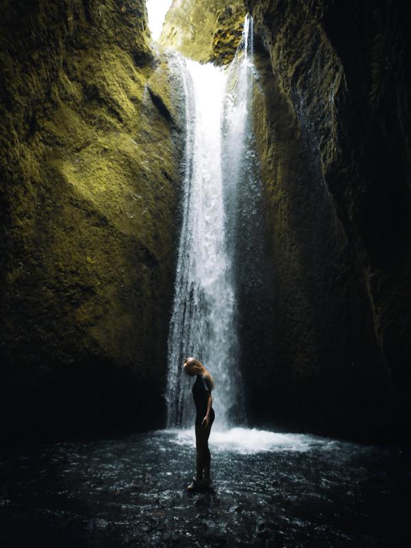 A woman stands in shallow water under a waterfall inside a mossy canyon.